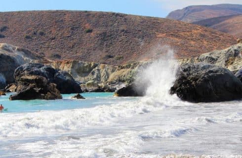 Waves crashing against dark rocks with a backdrop of hills under a clear blue sky.
