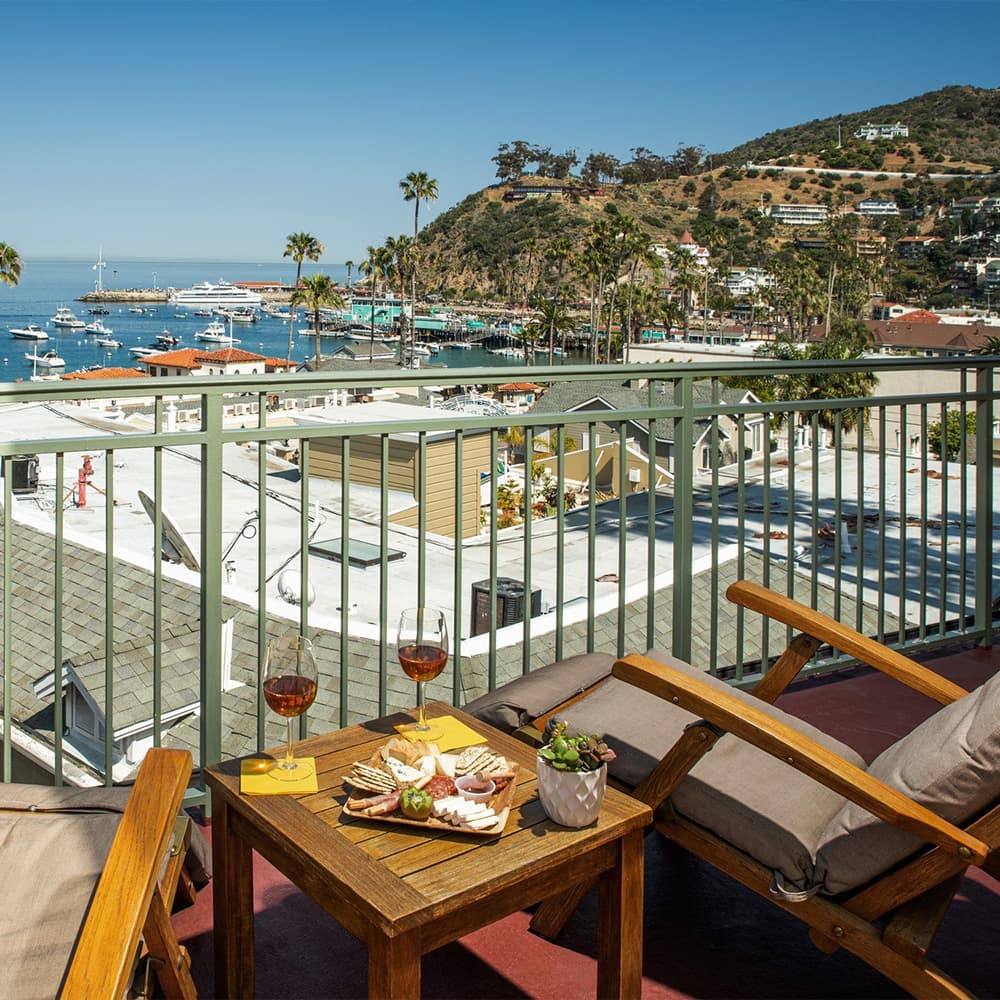 A coastal view from a balcony featuring two glasses of wine and a platter of snacks.