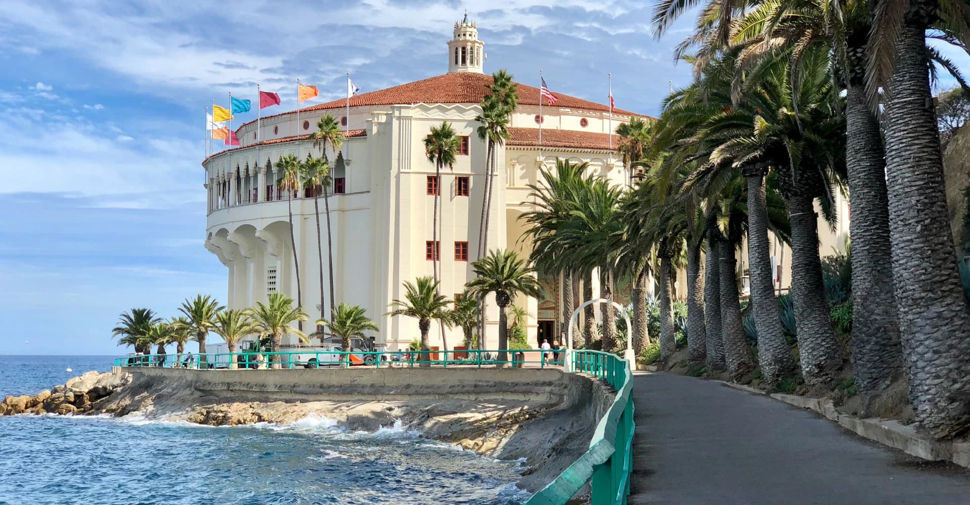 A seaside pathway lined with palm trees leads to a round, historic building with a red roof and flags.
